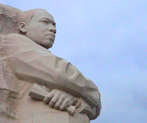 A towering stone sculpture shows a man with crossed arms holding a document, carved from a solid block against a cloudy sky.
