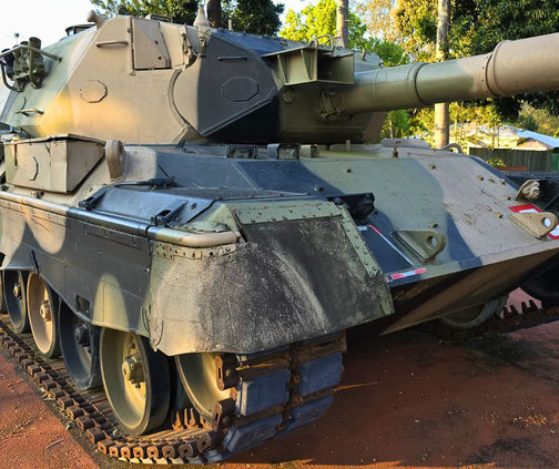 A close-up of a military tank with worn paint, heavy tracks, and a large barrel parked on a dirt surface.