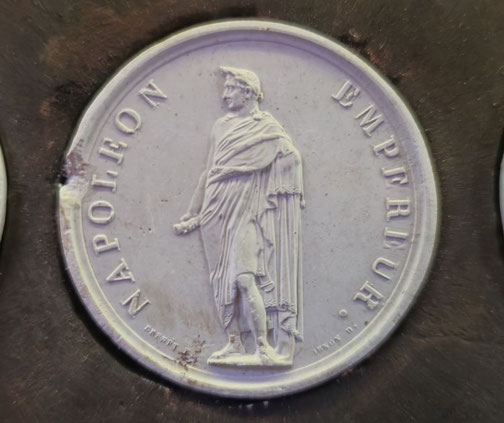 White medallion showing Napoleon I in a Roman-style robe, standing and facing slightly left, with "NAPOLEON" and "EMPEREUR" inscribed around the edge.