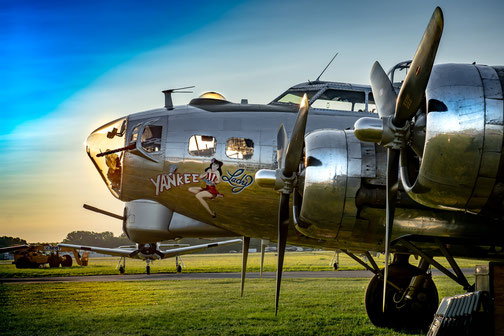 A polished B-17 bomber named "Yankee Lady" sits on a grassy airfield, featuring nose art of a woman and gleaming propellers at sunset.