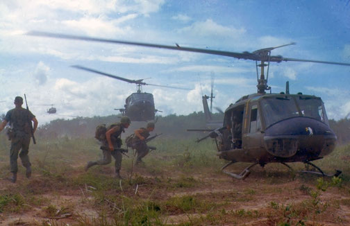 Soldiers disembark from helicopters in a grassy field during a military operation under a cloudy sky.