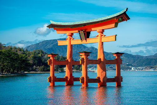 A large red torii gate stands in the water with mountains and forest in the background under a bright blue sky.
