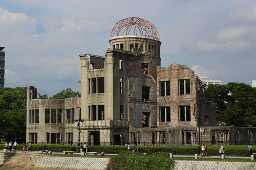 The Atomic Bomb Dome stands as a preserved ruin by a riverside walkway, with visitors nearby and trees in the background.