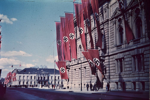 Large red Nazi flags with swastikas hang from a government building along a city street, with people walking below.
