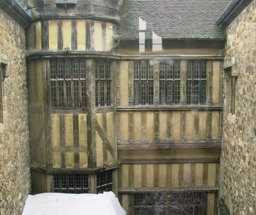 A historic timber-framed building with leaded glass windows and yellow plaster walls stands enclosed by stone walls. The structure showcases medieval architecture, featuring intricate wooden details.
