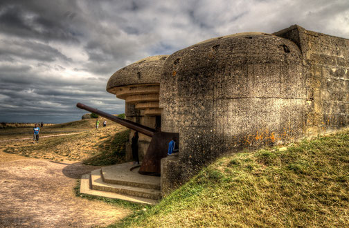 A large concrete World War II coastal bunker with an artillery gun faces an open landscape under cloudy skies.