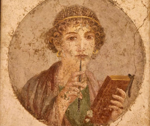 Ancient fresco of a woman with curly hair holding a stylus to her lips and a tablet in hand, suggesting thought and writing, often called the “Portrait of a Roman poetess.”