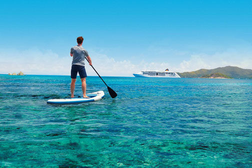 Person steht auf einem Stand-up-Paddle-Board im türkisblauen Meer, im Hintergrund die MS Europa 2 von Hapag-Lloyd Cruises.