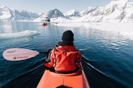 Person im Kajak auf arktischem Wasser mit Ponant-Paddel, im Hintergrund das Expeditionsschiff Le Commandant Charcot von Ponant, umgeben von Eisbergen und schneebedeckten Bergen.