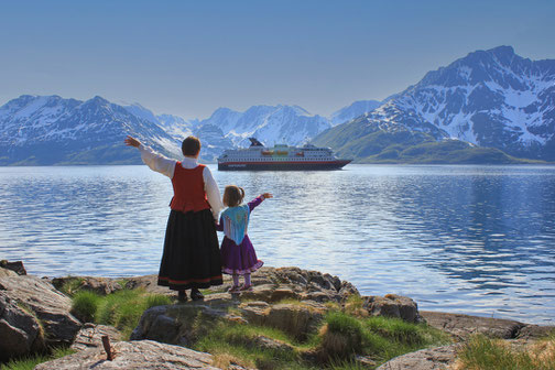 Frau in traditioneller Tracht und Kind blicken von der Küste auf ein Hurtigruten-Schiff inmitten norwegischer Fjorde.