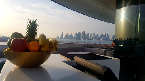 Oberdeck der Emerald Azzurra: Zu sehen ist eine Schale voller Früchte im Vordergrund und Ausblick auf Skyline von Doha im sanften Sonnenlicht.