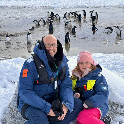 Timo Clarén von Emocean Cruises sitzt mit seiner Tochter an einem Strand in der Antarktis vor einer Gruppe Pinguine während einer Expedition mit der MS Fridtjof Nansen.