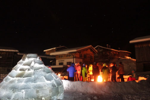 Schulklasse am Lagerfeuer neben beleuchtetem Iglu – Winter-Erlebnis-Klassenlager mit erlebnispädagogischen Aktivitäten