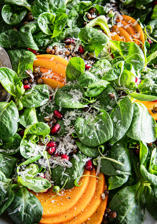 Feldsalat mit Beluga-Linsen, dünn geschnittenen Kaki-Scheiben, Granatapfelkernen und gehobeltem Parmesan auf einer Servierplatte, mit Balsamico-Dressing beträufelt.“