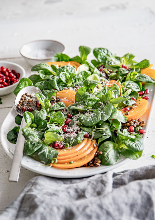 Feldsalat mit Beluga-Linsen, dünn geschnittenen Kaki-Scheiben, Granatapfelkernen und gehobeltem Parmesan auf einer Servierplatte, mit Balsamico-Dressing beträufelt.