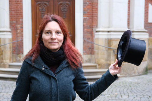 Frau mit roten Haaren, die einen Zylinder in der Hand hält.