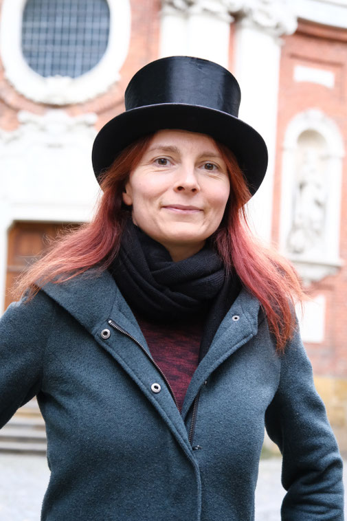 Frau mit Hut steht vor der Clemenskirche in Münster und lächelt.