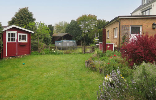 Gerade Rasenfläche vor der Terrasse, natürlich gewachsenes Beet, auf der Wiese steht ein rotes Gartenhaus
