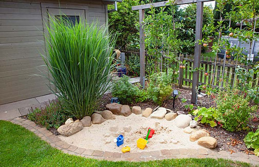 Ebenerdiger Sandkasten fügt sich durch seine geschwungene Umrandung in das Pflanzbeet ein