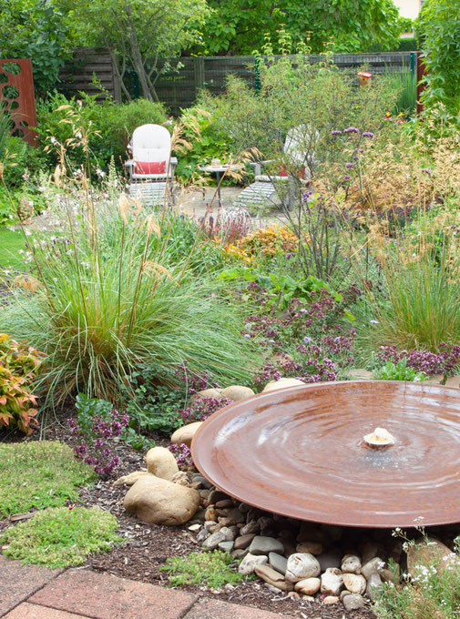 Runde Wasserschale aus Stahl ziert in einem Beet mit Steinen, Pflanzen und Blumen die Terrasse 