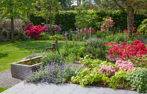 Steinbrunnen mit Schwengelpumpe ist vor Blumenbeet und Terrasse platziert