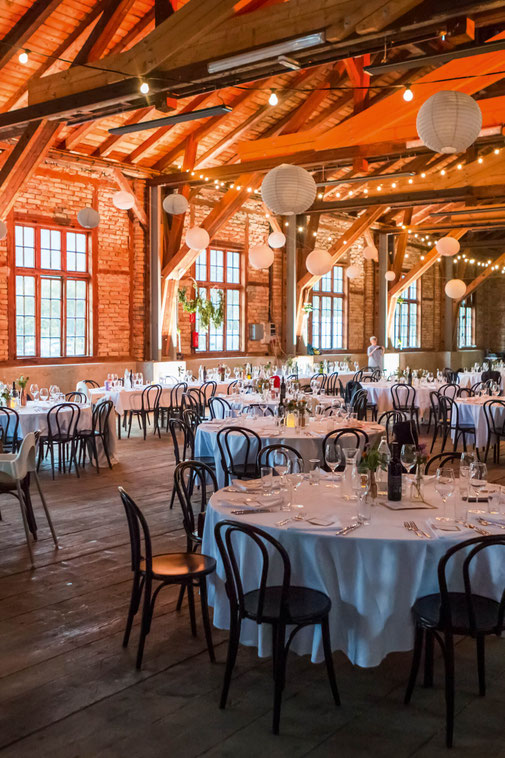 Festlich gedeckter Hochzeitsaal von Das Sägewerk mit runden Tischen, Lichterketten, Papierlampions, Backsteinwänden und Holzbalken.