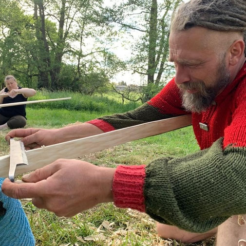 Mann bearbeitet draußen einen Holzbogen mit einem Ziehmesser.