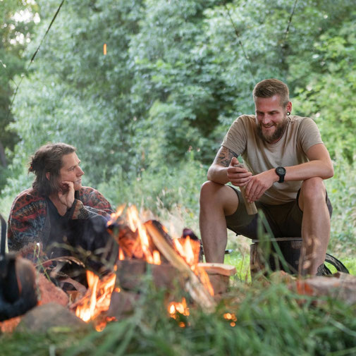 Zwei Menschen sitzen entspannt an einem Lagerfeuer im Grünen. 