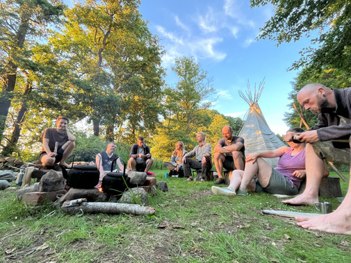 Gruppe sitzt im Kreis an einer Feuerstelle vor einem Tipi. 