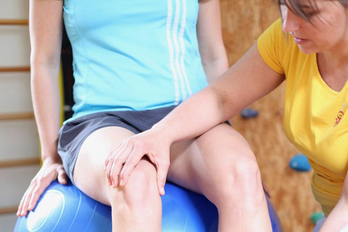 Physiotherapeutin korrigiert mit der Hand die Beinachse einer Patientin mit Knieschmerzen, die am Gynmastikball sitzt und eine Gleichgewichtsübung ausführt. Dies verbessert die Beschwerden am Knie vor.