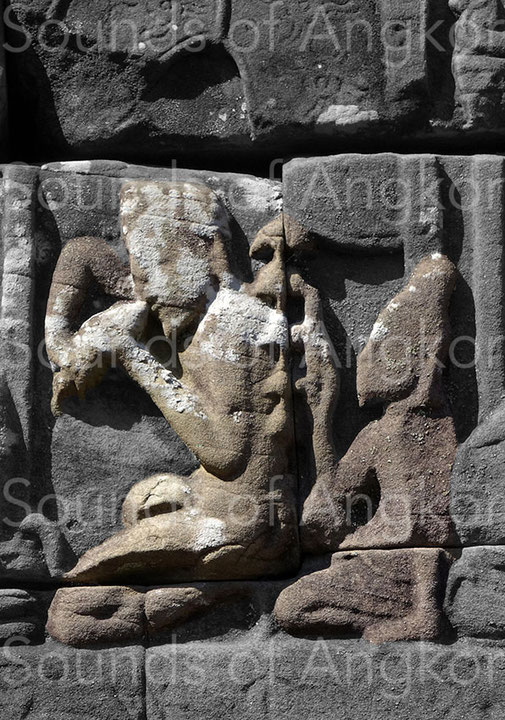 The left character blows into a conch while the one behind him hits a bell tree with five bells. Bayon. Exterior east gallery.