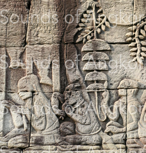 The second character from the left blows into a conch while the one behind him hits a bell tree with five bells. Bayon. Exterior gallery south.