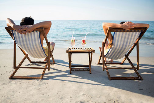 Zwei Urlauber sitzen mit nackten Füßen im Ostseesand, entspannt im Liegestuhl, dazwischen ein kleines Tischchen mit einem Drink