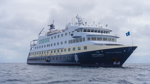MS Santa Cruz II fährt bei bewölktem Himmel durch ruhige See, mit Blick auf den Schiffsbug und die aufgezogene Flagge.