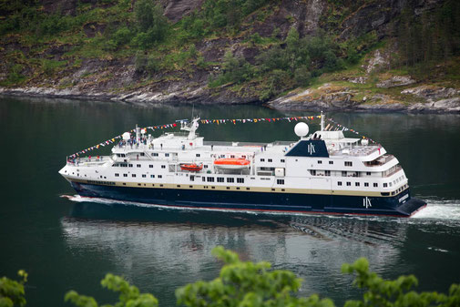 MS Spitsbergen mit Flaggen geschmückt, fährt durch einen grünen Fjord mit bewaldeten Berghängen im Hintergrund.