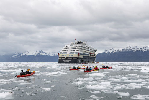 MS Roald Amundsen mit Kajaks im Eiswasser vor schneebedeckten Bergen.