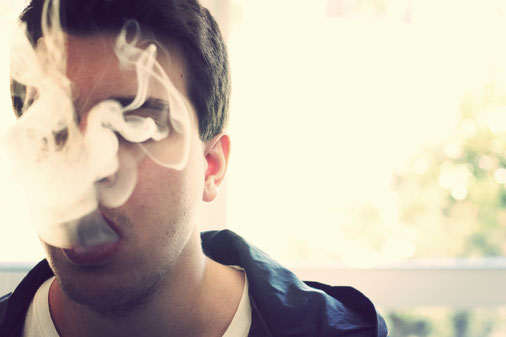 Ein mann mit einem blauen Kaputzenpulli raucht einenen Joint und atmet den Rauch langsam aus. Der Rauch steigt langsam vor sein Gesicht auf und verdeckt es.