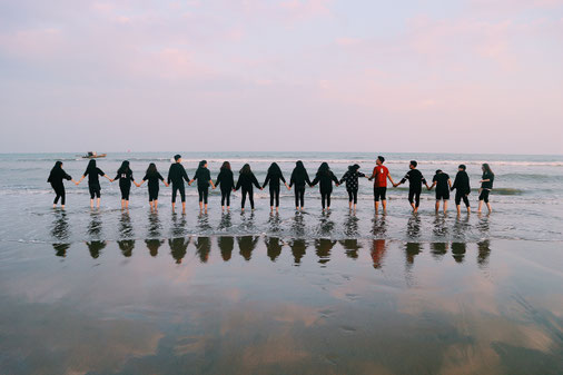 Gruppenbild am Meer, bei dem die 17 jungen Menschen durch an den Händen halten eine Kette bilden. Sie stehen barfuß im knöchelhohen Meer.ser