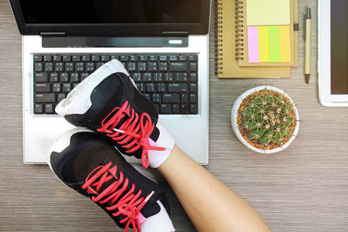 Beine mit Sportschuhen auf einem Schreibtisch mit Laptop und Arbeitsmaterialien