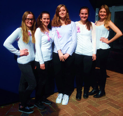 5 Mädchen, weiße T-Shirts, schwarze Hose, stehen vor blauem Hintergrund, rosa Schleife.