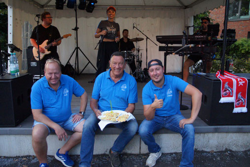 Drei Männer in bleiben T-Shirts sitzen am Rand einer Bühne und strahlen in die Kamera, eine Band spielt im Hintergrund.