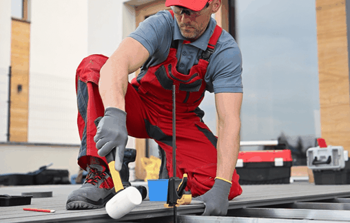 Handwerker in roter Arbeitskleidung montiert Bambus Terrassendielen auf einer Aluminium Unterkonstruktion mit einem weißen Gummihammer; im Hintergrund Werkzeugkoffer und modernes Gebäude
