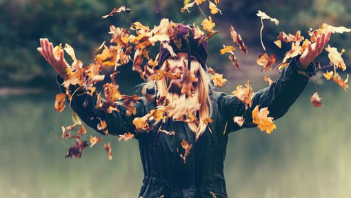 Frau im Herbstmantel wirft bunte Blätter in die Luft – Ausdruck von Freude, Leichtigkeit und Loslassen.“