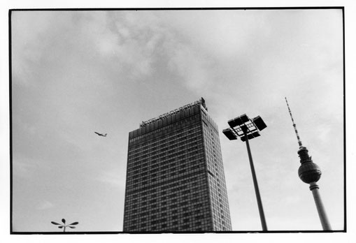 Ein FLugzeug fliegt vor dem Hotel Stadt Berlin, einer klassischen DDR-Straßenlaterne und dem Berliner Fernsehturm vorbei. Aufgenommen 1982.