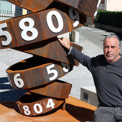 Paolo Grassi next to his conceptual public sculpture Thiago, featuring oxidized steel and white numbers in a spiral form.