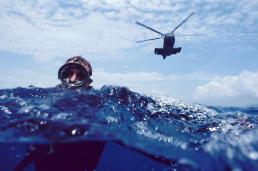 Pararescueman in the water; the rescue helicopter approaches slowly behind him.