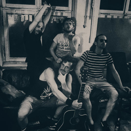 Die vier Mitglieder der BAnd in einem Übungsraum mit Akustikgitarre in Schwarz Weiß