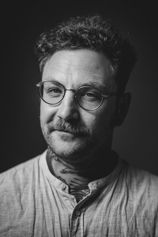 Tätowierter Mann mit Brille vor dunklem Hintergrund in Kamera blickend - Aufgenommen von Sebastian Schieder Porträt Fotograf Regensburg.