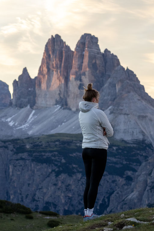 Eine Person in heller Outdoor-Kleidung steht auf einem felsigen Berggipfel neben einem schlichten Holzgipfelkreuz und blickt in die Weite der Dolomiten. Im Hintergrund färbt das sanfte Licht der Morgendämmerung den Himmel in zarten Rosa- und Blautönen und