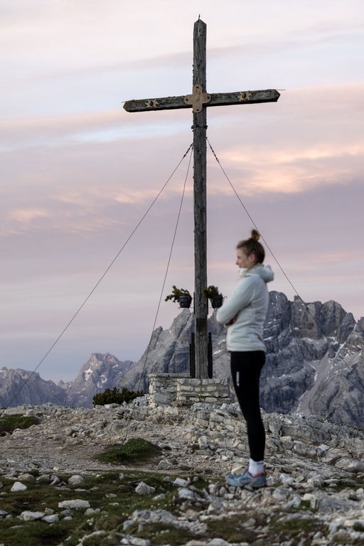 Die Drei Zinnen in den Dolomiten ragen markant in den Himmel und werden vom ersten warmen Licht der aufgehenden Sonne angestrahlt. Über den imposanten Felsformationen ziehen helle Wolken auf und verleihen der eindrucksvollen Berglandschaft eine ruhige, st